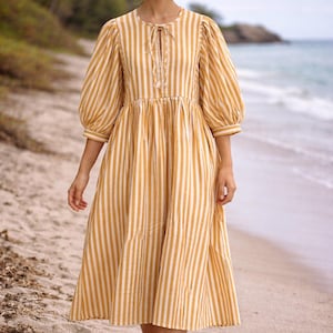 May include: A yellow and white striped dress with a tie-neck collar and elbow-length sleeves. The dress has a gathered waist and falls to mid-calf. The woman is walking on a sandy beach near the ocean. The dress is a casual, summery style.
