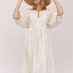 May include: A cream-colored linen dress with a tie-neck detail and voluminous sleeves. The dress is paired with a wide-brimmed straw hat with a yellow ribbon. The dress has a midi length and a gathered waist.