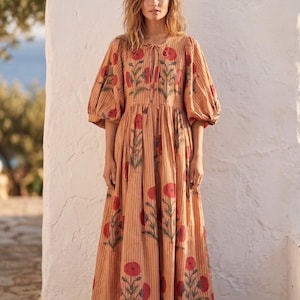 May include: A long, peach-colored dress with vertical stripes and red floral patterns. The dress features short, puffy sleeves and a tie at the neckline. The model is standing in front of a white wall.