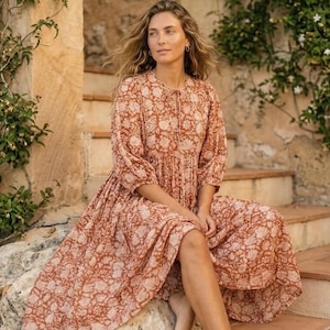 May include: A long, flowing dress with a floral pattern in shades of brown and cream. The dress has three-quarter sleeves and a relaxed fit. The model is sitting on stone steps, creating a bohemian aesthetic. The dress is perfect for a summer day.