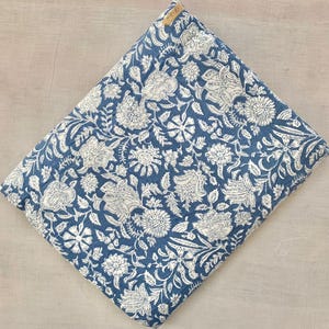 May include: A folded textile featuring a deep blue background with a dense, repeating white floral pattern. The fabric displays intricate botanical illustrations of various blossoms and leaves across the entire surface, with a small tan tag attached.