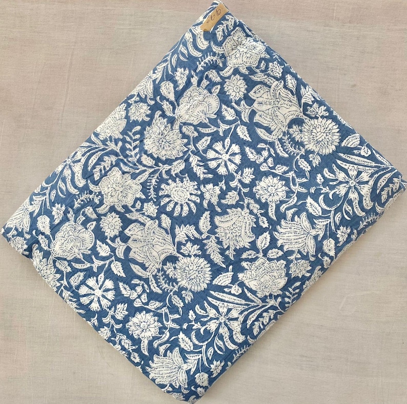 May include: A folded textile featuring a deep blue background with a dense, repeating white floral pattern. The fabric displays intricate botanical illustrations of various blossoms and leaves across the entire surface, with a small tan tag attached.