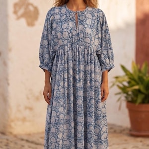 May include: A midi-length dress featuring a blue and white floral block print pattern. The garment has three-quarter length sleeves with gathered cuffs, a round neckline with a central tie closure, and a loose, flowing silhouette.