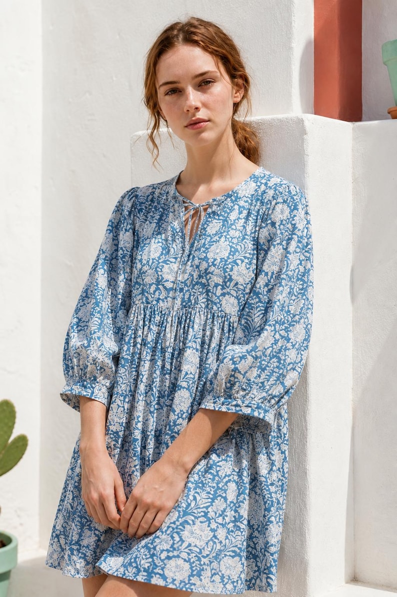 May include: A blue and white floral print dress featuring three-quarter length sleeves with gathered cuffs, a round neckline with a small tie closure, and a loose, flowing silhouette that falls above the knee with a gathered waistline detail.