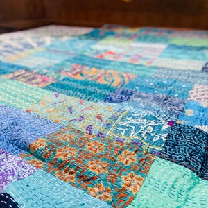 Puede incluir: Una colcha de retazos hecha de varias tonalidades de telas azules, verdes y moradas. La colcha está cosida con una puntada corrida, creando una superficie texturizada.