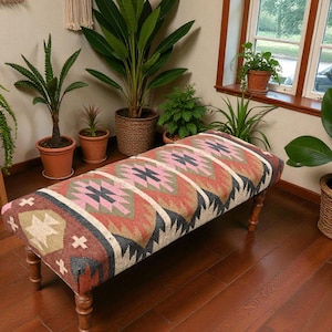 May include: A patterned bench with a rectangular seat and wooden legs. The seat displays a geometric design in pink, red, green, and black. The bench is set on a wooden floor, surrounded by plants and macrame wall decor.