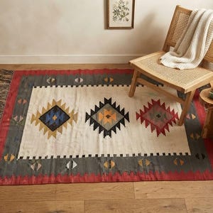 May include: A rectangular area rug featuring a cream-coloured centre with three geometric diamond patterns in blue, yellow, and red. The rug has a dark grey border with a red outer edge and fringe on the short ends, showcasing a woven textile design.