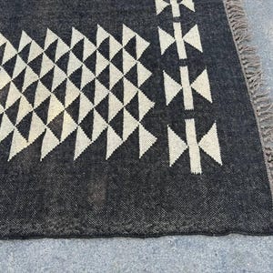 May include: A dark gray rug with a geometric pattern of white triangles and cross-like shapes. The rug has a textured surface and a fringed edge. The design is symmetrical and modern.