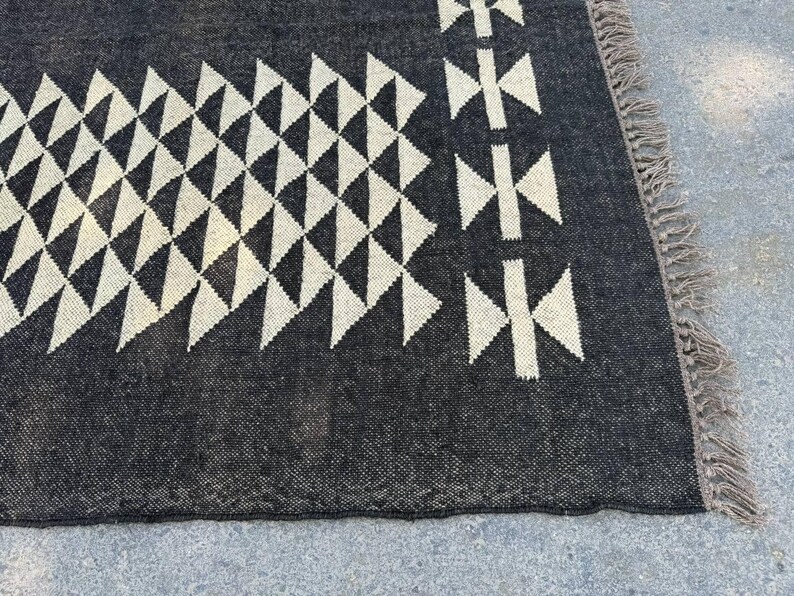 May include: A dark gray rug with a geometric pattern of white triangles and cross-like shapes. The rug has a textured surface and a fringed edge. The design is symmetrical and modern.