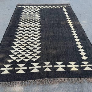 May include: A rectangular black rug with a geometric pattern of white triangles and diamond shapes. The rug has a fringe along the bottom edge. The rug is made of a woven material.