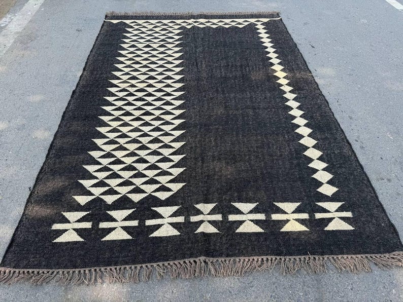 May include: A rectangular black rug with a geometric pattern of white triangles and diamond shapes. The rug has a fringe along the bottom edge. The rug is made of a woven material.