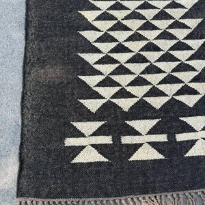 May include: A black and white geometric patterned rug. The rug features a repeating pattern of triangles and stylized shapes. The edges are finished with a fringe. The rug is made of a woven material.