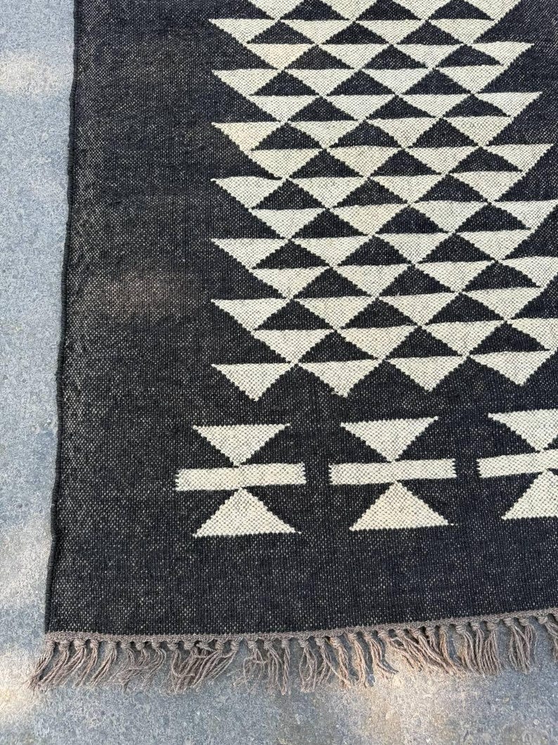 May include: A black and white geometric patterned rug. The rug features a repeating pattern of triangles and stylized shapes. The edges are finished with a fringe. The rug is made of a woven material.
