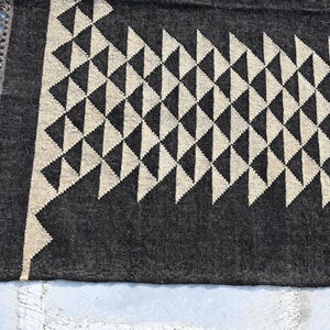 May include: A close-up of a dark gray rug with a repeating pattern of cream-colored triangles. The rug has a fringed edge and a geometric design, suitable for home decor.
