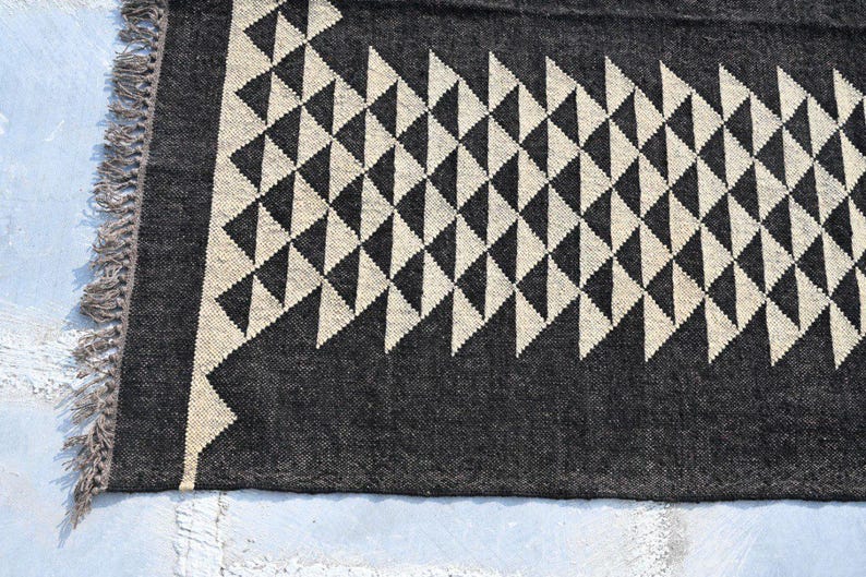 May include: A close-up of a dark gray rug with a repeating pattern of cream-colored triangles. The rug has a fringed edge and a geometric design, suitable for home decor.