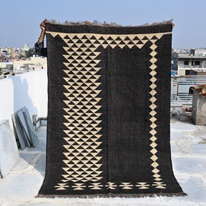 May include: A large, rectangular black rug with a geometric pattern of cream-colored triangles and diamonds. The rug is held up against a cityscape background, showcasing its design and texture. The rug has a fringed edge.