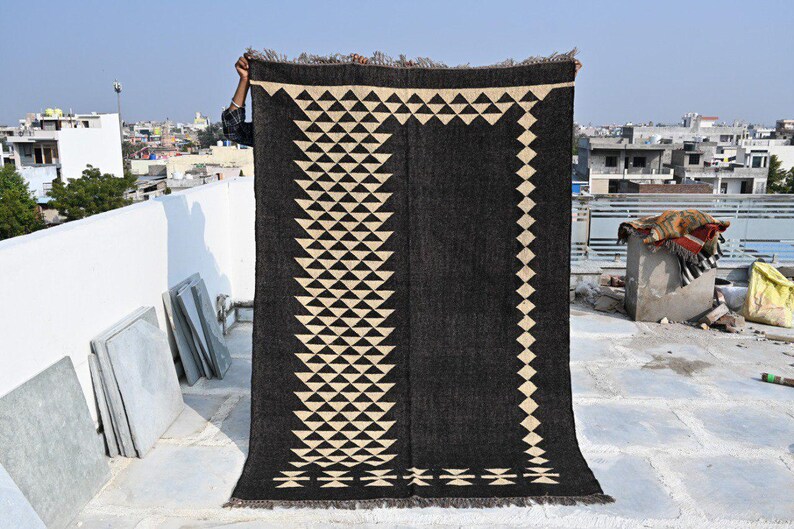 May include: A large, rectangular black rug with a geometric pattern of cream-colored triangles and diamonds. The rug is held up against a cityscape background, showcasing its design and texture. The rug has a fringed edge.