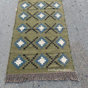 May include: Olive green rectangular rug with a geometric diamond pattern in white, black, and blue. The rug has a fringed edge and the text "JuteRugHandloom".