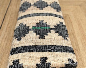 Puede incluir: Un banco rectangular con un diseño tejido. El banco presenta un fondo beige natural con patrones geométricos y rayas negras. Tiene cuatro patas de madera pequeñas y está colocado sobre una alfombra de yute.