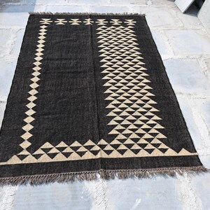 May include: A rectangular black rug with a geometric design. The rug features a border of diamond shapes and a central pattern of triangles in a beige color. The rug has a fringed edge.
