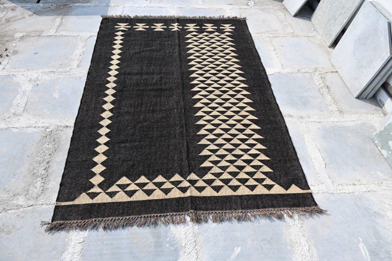 May include: A rectangular black rug with a geometric design. The rug features a border of diamond shapes and a central pattern of triangles in a beige color. The rug has a fringed edge.