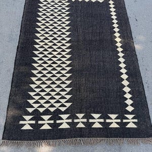 May include: A rectangular black rug with a geometric cream-colored pattern. The design features rows of triangles and diamond shapes, with a fringed edge. The rug is likely made of woven material and is suitable for indoor use.