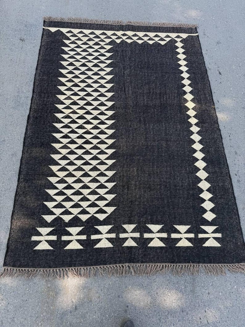 May include: A rectangular black rug with a geometric cream-colored pattern. The design features rows of triangles and diamond shapes, with a fringed edge. The rug is likely made of woven material and is suitable for indoor use.