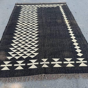 May include: A rectangular black rug with a geometric design. The rug features a pattern of white triangles and diamond shapes. The edges are finished with a fringe. The rug is suitable for indoor use.