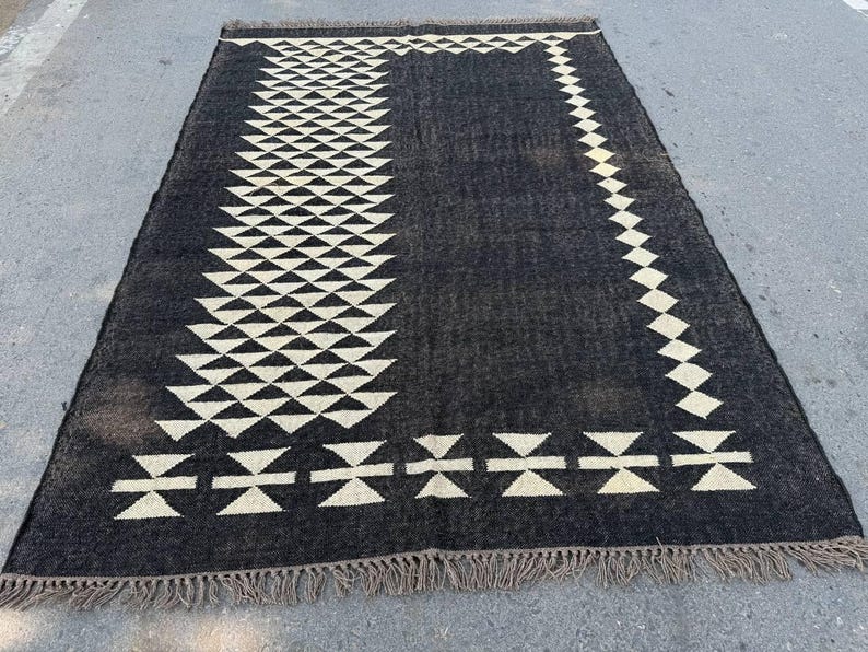 May include: A rectangular black rug with a geometric design. The rug features a pattern of white triangles and diamond shapes. The edges are finished with a fringe. The rug is suitable for indoor use.
