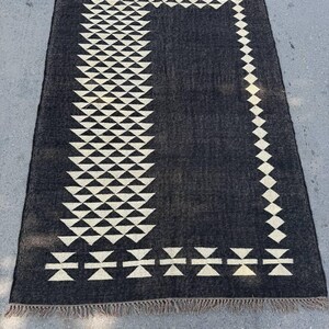 May include: A rectangular black rug with a geometric pattern. The rug features a repeating design of white triangles and diamond shapes along the border and a central column. The rug has a fringed edge.