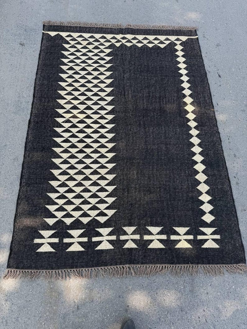 May include: A rectangular black rug with a geometric pattern. The rug features a repeating design of white triangles and diamond shapes along the border and a central column. The rug has a fringed edge.