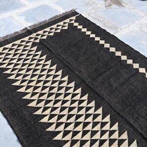 May include: A dark gray rug with a geometric design. The rug features a repeating pattern of cream-colored triangles and diamonds along the edges. The rug has a fringed edge and is made of a woven material.
