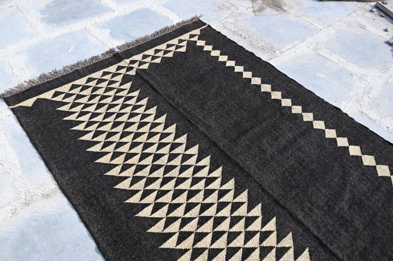 May include: A dark gray rug with a geometric design. The rug features a repeating pattern of cream-colored triangles and diamonds along the edges. The rug has a fringed edge and is made of a woven material.