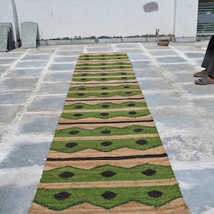 May include: A rectangular jute rug with a repeating pattern of green and tan stripes, accented with black diamond shapes. The rug is displayed outdoors on a concrete surface.
