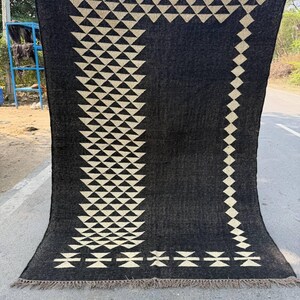 May include: A large, rectangular black rug with a geometric design. The rug features a repeating pattern of cream-colored triangles and diamonds. The edges are finished with a fringe. The rug is displayed outdoors.