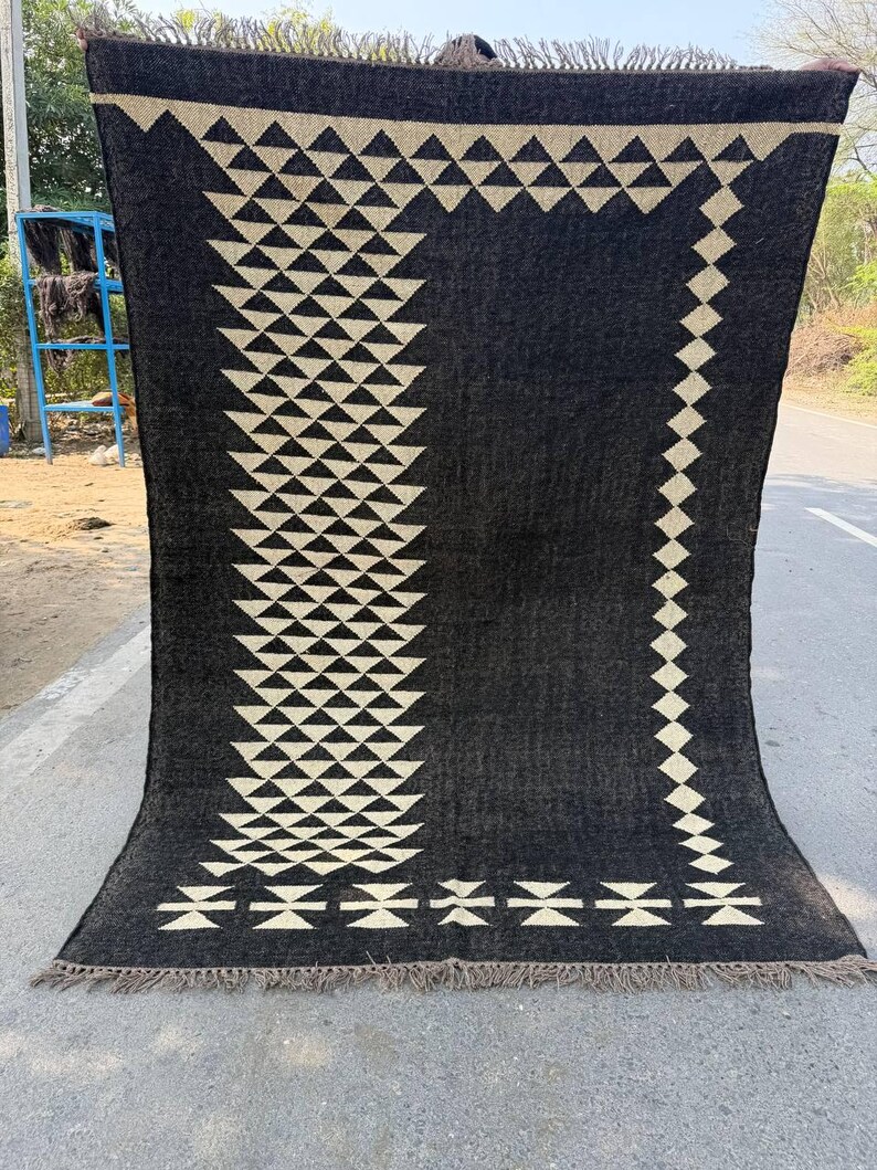 May include: A large, rectangular black rug with a geometric design. The rug features a repeating pattern of cream-colored triangles and diamonds. The edges are finished with a fringe. The rug is displayed outdoors.