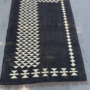 May include: A rectangular black rug with a geometric design. The rug features a border and a central vertical pattern of white triangles and diamond shapes. The rug has a fringed edge.