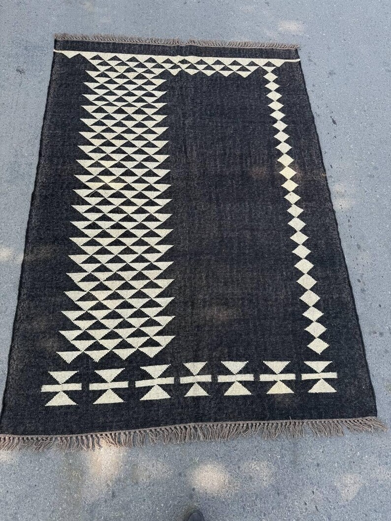May include: A rectangular black rug with a geometric design. The rug features a border and a central vertical pattern of white triangles and diamond shapes. The rug has a fringed edge.