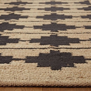 May include: A close-up of a woven rug with a geometric pattern in black and tan. The rug has a natural, rustic look and feel.