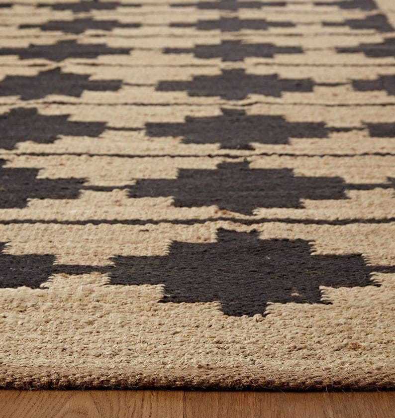 May include: A close-up of a woven rug with a geometric pattern in black and tan. The rug has a natural, rustic look and feel.