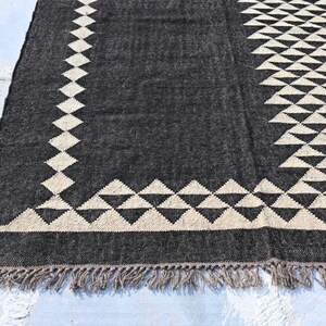 May include: A dark gray rug with a geometric design. The rug features a pattern of cream-colored diamonds and triangles along the edges and center. The rug has a fringe along the bottom edge.