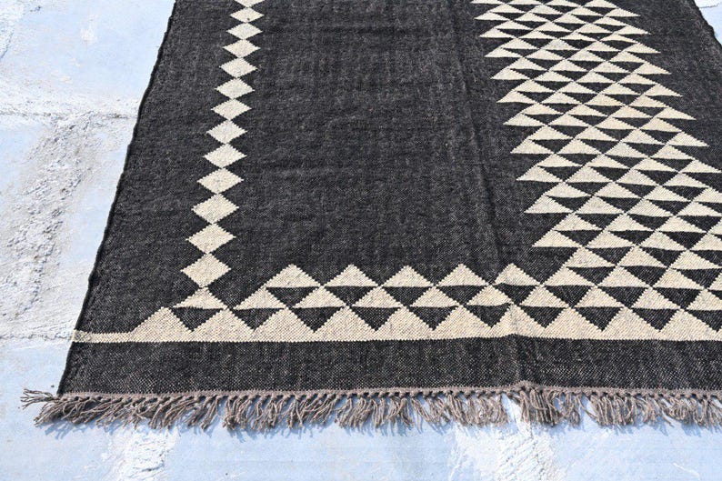 May include: A dark gray rug with a geometric design. The rug features a pattern of cream-colored diamonds and triangles along the edges and center. The rug has a fringe along the bottom edge.