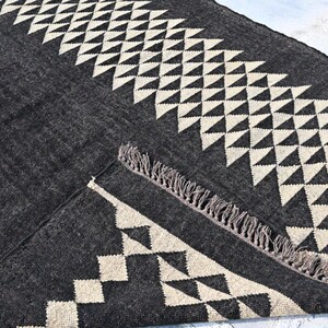 May include: A dark gray rug with a geometric pattern of white triangles and diamonds. The rug has a fringed edge and is made of a woven material. The design is symmetrical and modern, suitable for home decor.