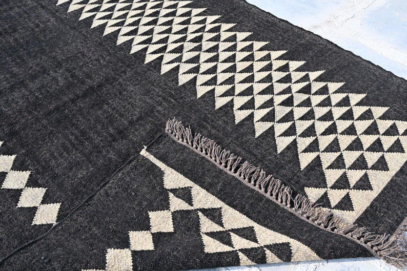 May include: A dark gray rug with a geometric pattern of white triangles and diamonds. The rug has a fringed edge and is made of a woven material. The design is symmetrical and modern, suitable for home decor.