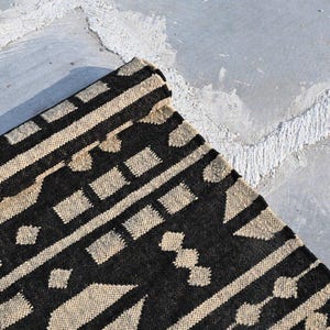 May include: A rectangular woven rug featuring a high-contrast pattern of black and beige geometric shapes. The design includes repeating horizontal bands of squares, diamonds, and linear stripes across the textured fabric surface.