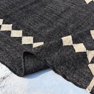 May include: A dark gray woven rug with geometric patterns. The rug features a repeating pattern of cream-colored diamonds along the border and triangles in the corner. The rug is folded over, revealing the texture and weave of the material.