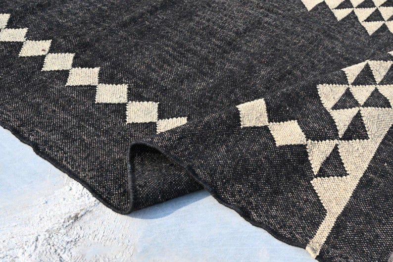 May include: A dark gray woven rug with geometric patterns. The rug features a repeating pattern of cream-colored diamonds along the border and triangles in the corner. The rug is folded over, revealing the texture and weave of the material.