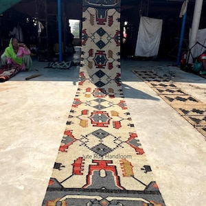 May include: A long, narrow jute rug with a geometric pattern in shades of red, blue, yellow, and white. The rug is handloom and has the text "Jute Rug Handloom" printed on it.