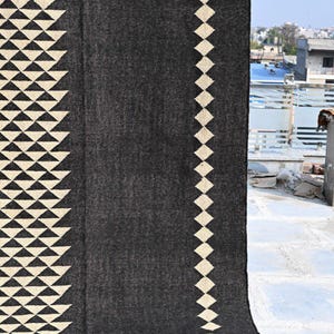 May include: A dark gray rug with geometric patterns. The rug features cream-colored triangles along the edges and a diamond pattern on one side. The bottom edge has a fringed detail. This rug is suitable for home decor.