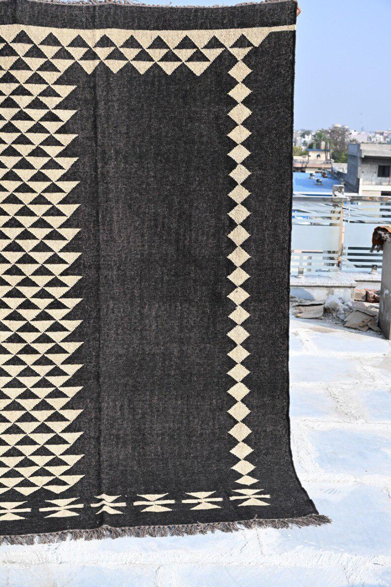 May include: A dark gray rug with geometric patterns. The rug features cream-colored triangles along the edges and a diamond pattern on one side. The bottom edge has a fringed detail. This rug is suitable for home decor.